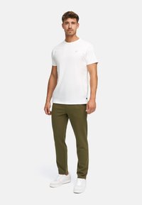 Mand, der står og har en ensfarvet hvid kortærmet t-shirt, olivenfarvede bukser og hvide sneakers på en lys grå baggrund.