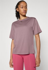 Femeie îmbrăcată într-un tricou scurt din material mauve de la Puma și leggings roz de la Puma, stând în fața unui fundal simplu deschis și privind în jos.