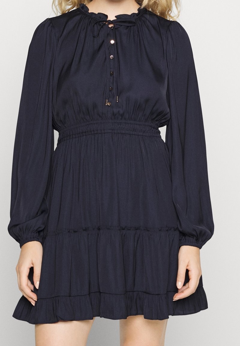 Femme portant une robe noire à manches longues avec des boutons, un col à volants, une taille élastique et une jupe à étages, debout devant un fond uni.
