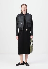 Μαύρο μπουφάν bomber με γυαλιστερή ύφανση, δύο μπροστινές τσέπες και χρυσά αξεσουάρ, σε συνδυασμό με ένα μακρύ μαύρο φόρεμα και πράσινη τσάντα. Επιλέγονται επίπεδες παπούτσια.