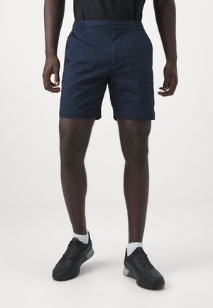 Männliches Modell trägt dunkelblaue Shorts, schwarze Sneaker mit weißen Socken, steht vor einem einfachen hellen Hintergrund.