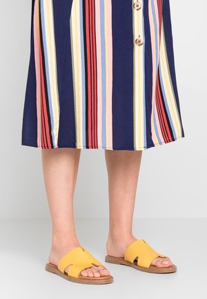 Gule slidesandal med udskåret design, korksål og struktureret overdel, kombineret med en knælængde kjole med striber i marineblå og pastelfarver.