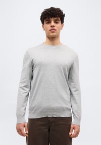 Jeune homme aux cheveux bouclés portant un pull léger à manches longues gris clair et un pantalon marron foncé, debout devant un fond clair uni.