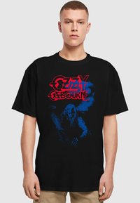 Sort bomulds t-shirt med blå og rød grafisk tryk af "Ozzy Osbourne" tekst og en illustreret figur mod en mørk baggrund.