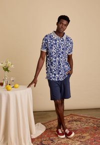 Blauw-wit gestreepte polo met een lichtgrijze kraag, gecombineerd met marineblauwe shorts en rode sneakers. Eenvoudige achtergrond met een tafel en citroenen.