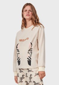 Sudadera color crema con cuello redondo, que presenta un diseño en la parte frontal de caras de cebra en negro y rosa, y las palabras "WILD WILD" arriba.