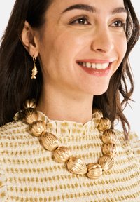 Collier en or avec des perles arrondies et côtelées ; boucles d'oreilles en or en forme de poisson assorties ; porté avec un haut blanc texturé et à volants avec des accents jaunes.