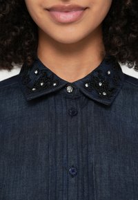 Rifľová košeľa tmavomodrej farby s čiernym vzorovaným vyšívaným golierom ozdobeným priehľadnými korálkami a dekoratívnym gombíkom.