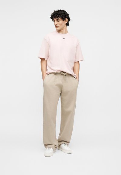 Νεαρός άνδρας με σγουρά μαλλιά, φοράει ροζ ανοιχτό oversized T-shirt, μπεζ φαρδιά παντελόνια, λευκά αθλητικά παπούτσια, στέκεται με τα χέρια στις τσέπες.