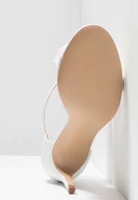 Chaussure à talon haut blanche au design élégant, matériau synthétique lisse, bout pointu et semelle beige texturée. Équipée d'une bride à la cheville.
