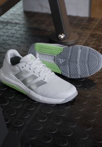 Chaussures de sport blanches avec des rayures grises et des accents verts sur un sol de gym noir texturé, une chaussure montrant le dessus et l'autre la semelle.
