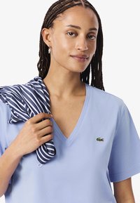 Donna con capelli intrecciati che indossa una maglietta azzurra a scollo a V con logo di coccodrillo, mentre tiene una stoffa a righe blu navy e bianca sulla spalla.