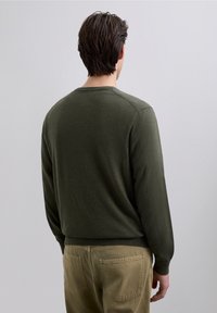 Suéter verde oscuro con cuello redondo, mangas largas y puños acanalados. Textura suave con diseño entallado, combinado con pantalones caqui claros.
