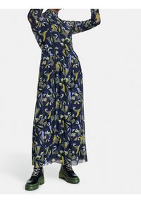 Robe maxi sur fond marine avec des motifs floraux en vert, jaune et violet. Dotée de manches longues et d'un col montant, accompagnée de bottes noires.