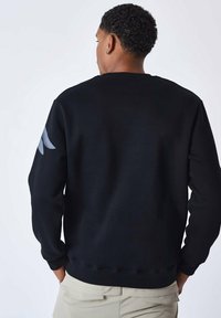 Schwarzer Pullover aus Baumwollmischung mit Rundhalsausschnitt und langen Ärmeln. Ein Ärmel zeigt ein grafisches graues Detail. Minimalistisches Design.