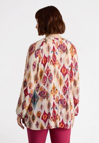 Långärmad blus i lätt tyg med ett mönster av flerfärgade diamanter och blommor på en krämfärgad bakgrund.