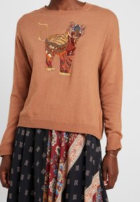 Pull en maille marron avec un motif de lion coloré brodé. Le lion présente des motifs et des textures complexes, créant un élément visuel vibrant.