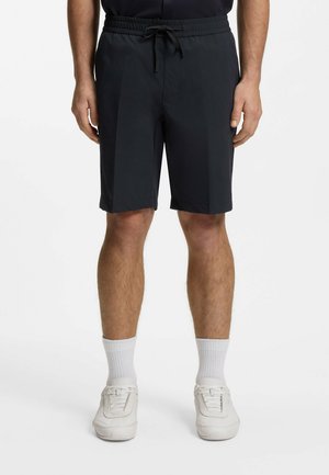 Mężczyzna noszący czarne szorty do kolan z troczkiem, białe skarpetki za kostkę oraz białe trampki, stojący na białym tle.