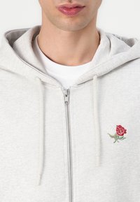 Lys grå zip-up hettegenser laget av myk stoff, med broderi av en hvit og rød rose på venstre brystområde og snorer ved hetten.