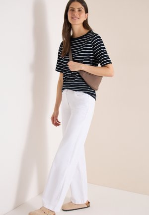 Vrouw die witte wijde broek, zwart-wit gestreept kort shirt, beige schoudertas en beige instapschoenen draagt, staand binnen.