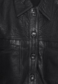 Sort læder skjorte med tekstureret overflade, stor krave og knaplukning foran. Har to lommer og blanke sorte knapper.