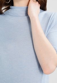 Ljusblå topp med högt krage och korta ärmar, mjuk textur och figursydd silhuett, med ett slätt, lätt genomskinligt tyg.