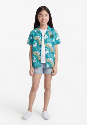 Νεαρή κοπέλα στέκεται, φορώντας μια μπλούζα με κοντά μανίκια σε τιρκουάζ με σχέδια πάνω από ένα λευκό μπλουζάκι, ανοιχτόχρωμα τζιν σορτς και άσπρα παπούτσια χωρίς κορδόνια.