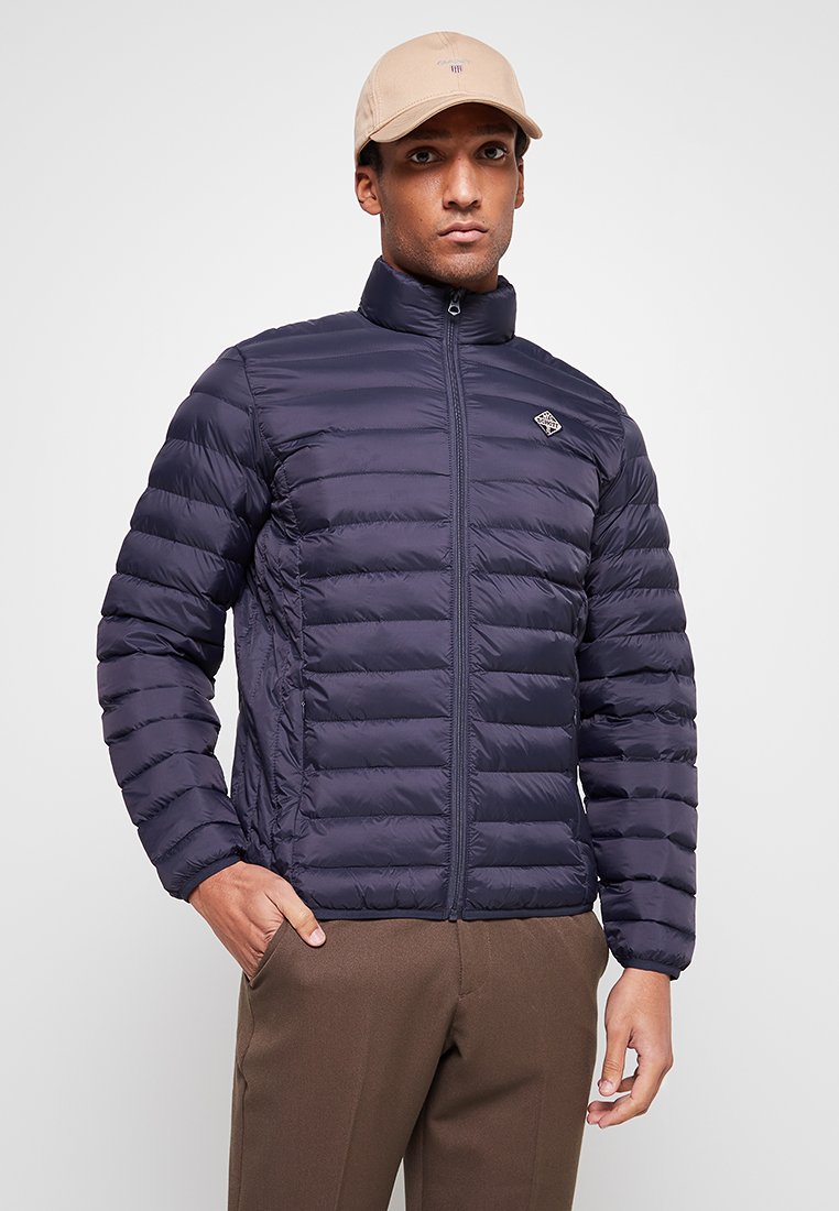 Mann, der eine marineblaue Steppjacke, eine beigefarbene Mütze und braune Hosen trägt, mit einer Hand in der Tasche vor einem schlichten hellen Hintergrund steht.