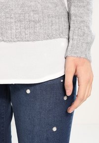 Grijze gebreide trui over een witte top, gecombineerd met blauwe jeans met parelaccenten en een hand die op het been rust.