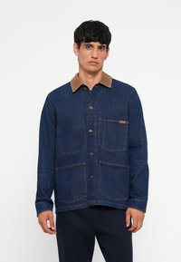 Chaqueta de denim azul oscuro con un cuello de pana color marrón claro, dos grandes bolsillos frontales y cierre de botones, con un ajuste limpio y entallado.