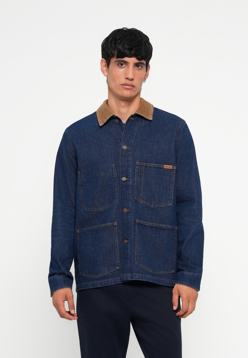 Chaqueta de denim azul oscuro con un cuello de pana color marrón claro, dos grandes bolsillos frontales y cierre de botones, con un ajuste limpio y entallado.