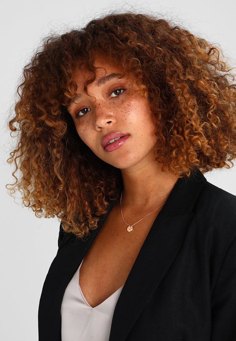 Cheveux bruns bouclés, peau tachetée, portant un blazer noir sur un haut clair. Présente un collier délicat et une couleur de lèvres naturelle.