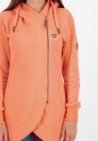 Haut à zip orange avec capuche, poches avant et logo en forme de cœur. Dispose de poignets côtelés et d'un ourlet asymétrique. Texture de tissu lisse.