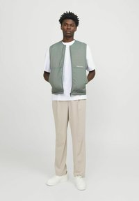 Groene gevoerde vest met ritssluiting, voorzien van een logo op de borst, gedragen over een wit T-shirt en gecombineerd met beige broek en witte sneakers.