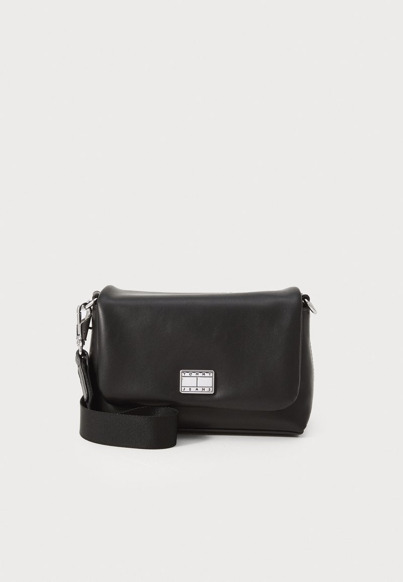 Bolso cruzado de cuero negro con cierre de solapa magnético, correa de tela ajustable y una placa de logo plateado en el frente.