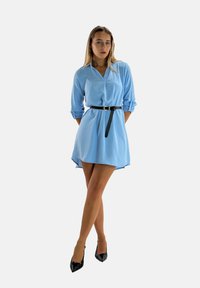 Light blue shirt dress with rolled sleeves, a v-neckline, and a high-low hem, cinched at the waist with a black belt. Black pointed heels.