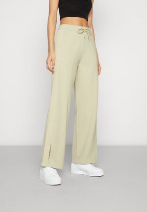 Kvinde iført beige vidde bukser med forreste slidser, snøre i taljen, sort kort top og hvide sneakers mod en ensfarvet baggrund.