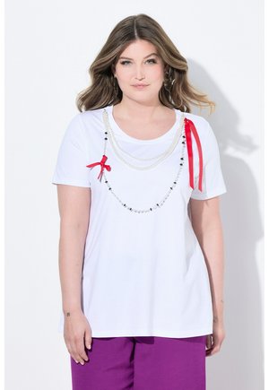 Frau mit welligem Haar trägt ein weißes T-Shirt, das mit mehrlagigen Perlen- und schwarzen Perlenketten mit roten Bändern dekoriert ist, kombiniert mit lila Hosen.