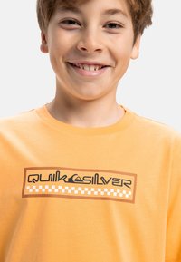 Oranžové bavlnené tričko s čiernym a hnedým logom Quiksilver, obohnané kockovaným vzorom na spodnej časti. Krátke rukávy, okrúhly výstrih.