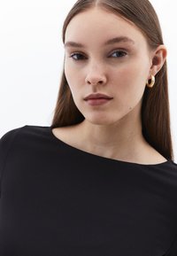 Schwarzes Stoffoberteil mit weitem Ausschnitt, kombiniert mit goldenen Creolen. Glatte Textur, minimalistisches Design, keine Muster oder auffälligen Akzente.