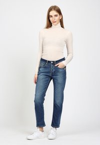 Helles beige Langarm-Turtleneck-Oberteil, kombiniert mit mittel-hoch geschnittenen blauen Jeans und weißen Sneakers. Bemerkenswerte Details sind die taillierte Silhouette und die leichte Textur.