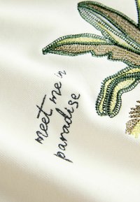 Vyšívané zelené a žlté lístky s vyšitou frázou „stretneme sa v raji“ na bielom látke.