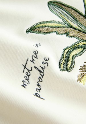 Valgel riidel tikitud rohelised ja kollased lehed ning tikandiga fraas "kohtume paradiisis".