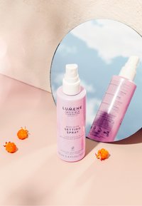 Fioletowa butelka sprayu z białą pompką, opisana jako "Spray Utrwalający z Efektem Rosy", ustawiona na różowym tle i odbijająca się w okrągłym lustrze.