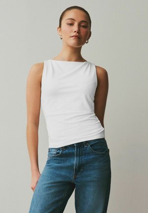 Junge Frau mit zurückgebundenen Haaren, trägt ein ärmelloses weißes Oberteil und blaue Jeans, steht vor einem schlichten hellen Hintergrund.