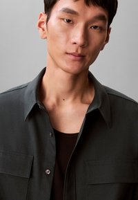 Camicia verde scuro con colletto, maniche lunghe, due tasche frontali e chiusura con bottoni; indossata sopra un top a costine scuro. Tessuto strutturato.