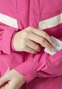 Lesklá růžová bunda s bílými pruhy, s zipovým zapínáním a kapsou na rukávu. Hladký materiál a přiléhavé manžety. Karta, která je vložena do kapsy.