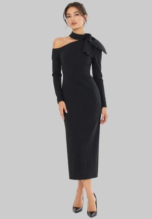 Frau trägt ein eng anliegendes schwarzes Midikleid mit einer freiliegenden Schulter und einer großen Schleife in der Nähe des Halses, kombiniert mit schwarzen High Heels.