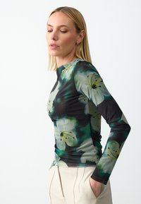 Femme aux cheveux blonds lisses portant un haut ajusté à longues manches avec un grand imprimé floral blanc et un pantalon crème, la main dans la poche.