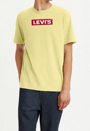 Hombre con camiseta de color amarillo claro con el logo de Levi's y jeans azul oscuro, de pie con una mano en el bolsillo contra un fondo liso.